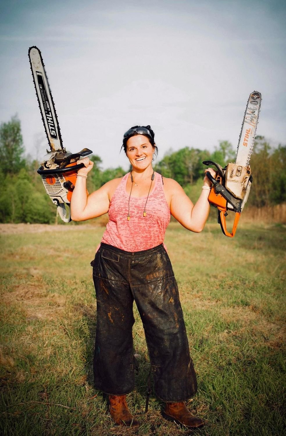 A carver stands in a field holding a chainsaw in each hand.